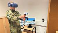 Ein Soldat steht in einem Raum. Er trägt eine VR-Brille und hat VR-Steuerungstools in beiden Händen.