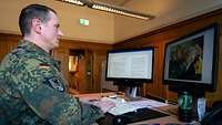 Ein Soldat sitzt an einem Schreibtisch vor zwei Bildschirmen und tippt etwas auf der Tastatur