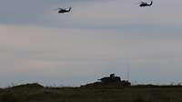 Ein Schützenpanzer Puma steht bei Dämmerung auf einem Hügel. Im Hintergrund fliegen zwei Hubschrauber.