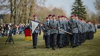 Soldaten marschieren in Formation über eine Wiese, vorn eine Soldatin mit weißer Dreiecksfahne