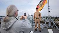 Ein Marinesoldat in sandfarbener Uniform steht am Heck eines Schiffes und wird mit Handy fotografiert.