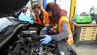Two girls wearing blue protective gloves looking inside the engine bay of a passenger car. 