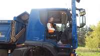 A young woman sits on the passenger seat of a blue dump truck with the door open.