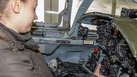 A young woman sitting in the cockpit of a fighter aircraft holding the yoke in her right hand.