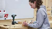 A young woman standing at a workbench drawing a plane across a clamped piece of wood.