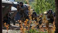 Mehrere Soldaten feuern mit ihrem Gewehr neben Militärfahrzeugen, im Vordergrund Pflanzen.