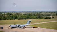 Ein militärisches Transportflugzeug steht am Boden, ein weiteres befindet sich im Landeanflug.