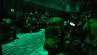 In einem Flugzeug sitzen bei grünem Licht Soldaten mit riesigen Rucksäcken und Fallschirmen.