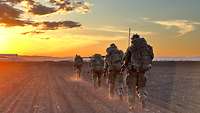 Vier Soldaten laufen hintereinander über ein Feld. Die Sonne geht unter, der Himmel leuchtet gelb.