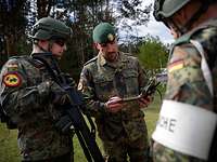 Ein Soldat im Gelände spricht und schaut auf seine Notizen, zwei bewaffnete Soldaten hören ihm zu. 