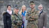 Eine Soldatin, ein Soldat und drei Zivilisten dahinter posieren für ein Gruppenbild