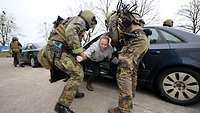 Zwei Soldaten in voller Montur ziehen eine Frau aus einem Auto und bringen sie zu Boden