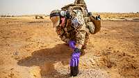 Ein Soldat im Wüstentarn kniet in der Wüste mit einem schwarzen Behälter vor einer Kuhle