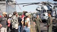 Menschen stehen auf dem Flugdeck eines Schiffes, ein Soldat in olivgrünem Overall spricht mit ihnen.