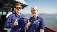 A man and a woman in blue work uniforms on the deck of a ship show a thumbs up.