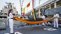 Four sailors in white uniforms hold a large falg coloured black, red and gold.