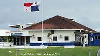 Vor einem weiß-blauen Haus weht die Panama Flagge, im Grass der Schriftzug Canal de Panama.