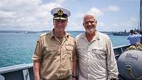 Ein Mann in Uniform und ein Mann in Zivil, stehen nebeneinander auf einem Schiff.