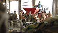 Soldaten in einer Halle beim Aufbau der Sandsack-Füllmaschine, im Hintergrund ein Helfer des THW.