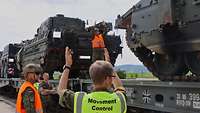 Zwei Soldaten zeigen per Handzeichen einen Abstand an, einer steht auf einem Waggon mit Schützenpanzern