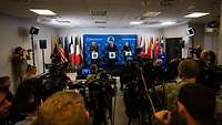 Three men stand in a room at lecterns in front of media representatives sitting opposite them.