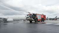 Ein Feuerwehrlöschfahrzeug steht vor Flugzeugen und sprüht aus zwei Düsen im hohen Bogen Wasser. 