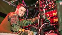 Ein Soldat posiert vor der Rückseite eines IT-Geräts mit zahlreichen angesteckten Kabeln fürs Foto 