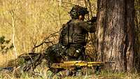 Zwei Soldaten hocken und liegen mit Gewehr im Anschlag hinter einem Baum in Deckung