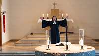Frau im Pfarrersgewand vor Altar hebt die Hände und spricht. Vor ihr eine Bibel, Kerzen und Kreuz.