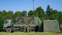 Soldaten stehen vor einem Laster mit Flecktarn-Aufbau, daneben ein grünes Zelt.