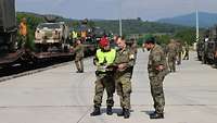 Drei Soldaten stehen auf einem Bahngelände und unterhalten sich, im Hintergrund mehrere Waggons mit Militärfahrzeugen 