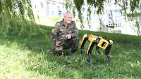 A man in uniform is kneeling on a meadow next to a yellow-black robot on four legs.