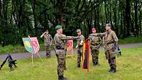 Mehrere Soldaten stehen auf einer Wiese. Neben ihnen sind die Truppenfahne, zwei Wappen und zwei Waffenpyramiden.