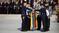 Soldaten stehen auf einem Platz vor Zuschauern und legen ihre Hand über die deutsche Flagge