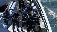 Marinesoldaten in blauer Arbeitsuniform und Schutzhelm auf dem Oberdeck eines Schiffes.