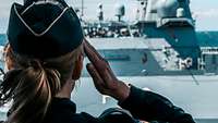 Eine Marinesoldatin mit blauem Schiffchen grüßt militärisch ein graues Kriegsschiff. 