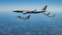Drei Kampfjets begleiten ein großes Flugzeug am Himmel