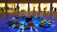 Blumenkränze liegen in einem Halbkreis in einer unterirdischen Gedenkstätte
