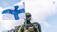 Ein Soldat in Gefechtsanzug mit finnischer Flagge vor blau-weißem Himmel