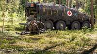 Drei Soldaten knien im Wald am Boden um eine liegende Person, hinter ihnen ein Sanitätsfahrzeug