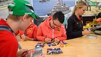 Drei Frauen in Sportanzügen signieren an einem halbrunden Counter Autogrammkarten
