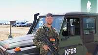 Ein Soldat steht mit einem Gewehr vor einem Einsatzfahrzeug und posiert in die Kamera