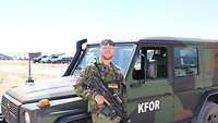 Ein Soldat steht mit einem Gewehr vor einem Einsatzfahrzeug und posiert in die Kamera