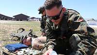 Ein Soldat liegt verbunden am Boden. Ein Soldat im Vordergrund bereitet Verbandsmaterial vor.