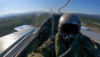 A pilot sits in the cockpit during a flight over a landscape.