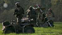 Drei Soldaten in Kampfausrüstung stehen mit militärischen Fahrzeugen auf einem Feld