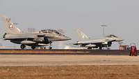 Two fighter jets stand on the airfield with their engines running.