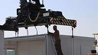 Ein Soldat befestigt auf einer Leiter stehend den Container mit Ketten am Tragegestellt des Hysters