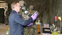 Ein Mann im Anzug und ein Soldat mit ABC-Schutzmaske halten ein Laborgerät in den Händen und blicken darauf