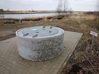 ein Betonzylinder steht auf einer gepflasterten Fläche auf einer Wiesenfläche neben einem Gewässer.